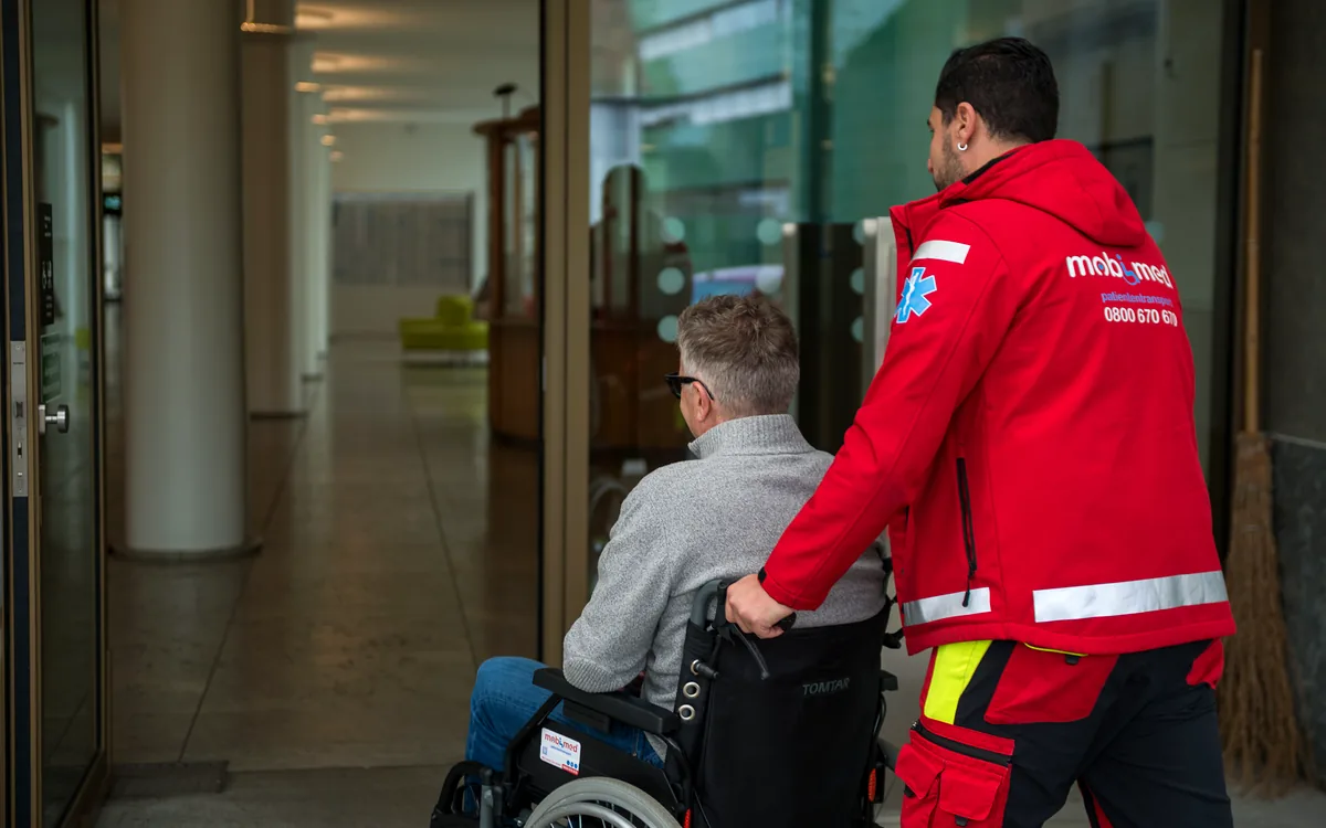 Mobimed-Mitarbeiter begleitet einen älteren Herrn im Rollstuhl in die Eingangshalle