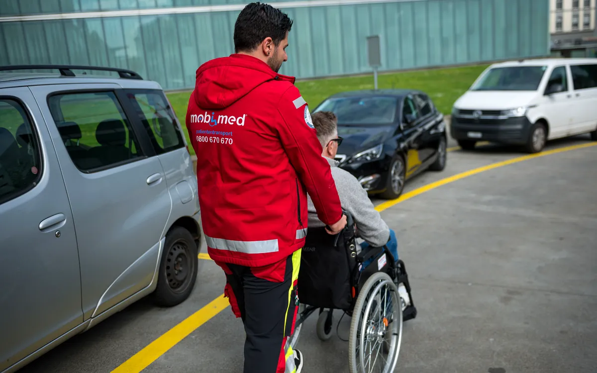 Mobimed-Mitarbeiter schiebt einen Patienten im Rollstuhl zum Fahrzeug