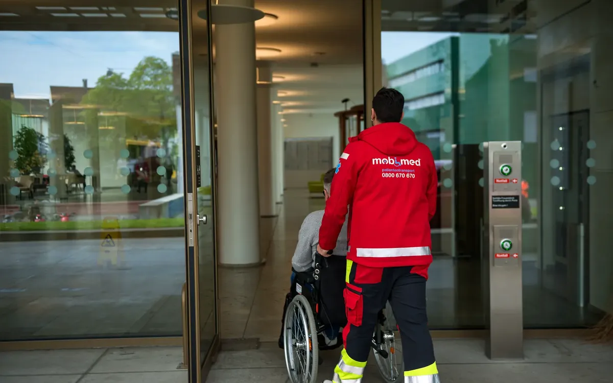 Mobimed-Mitarbeiter begleitet einen Patienten im Rollstuhl in ein Gebäude