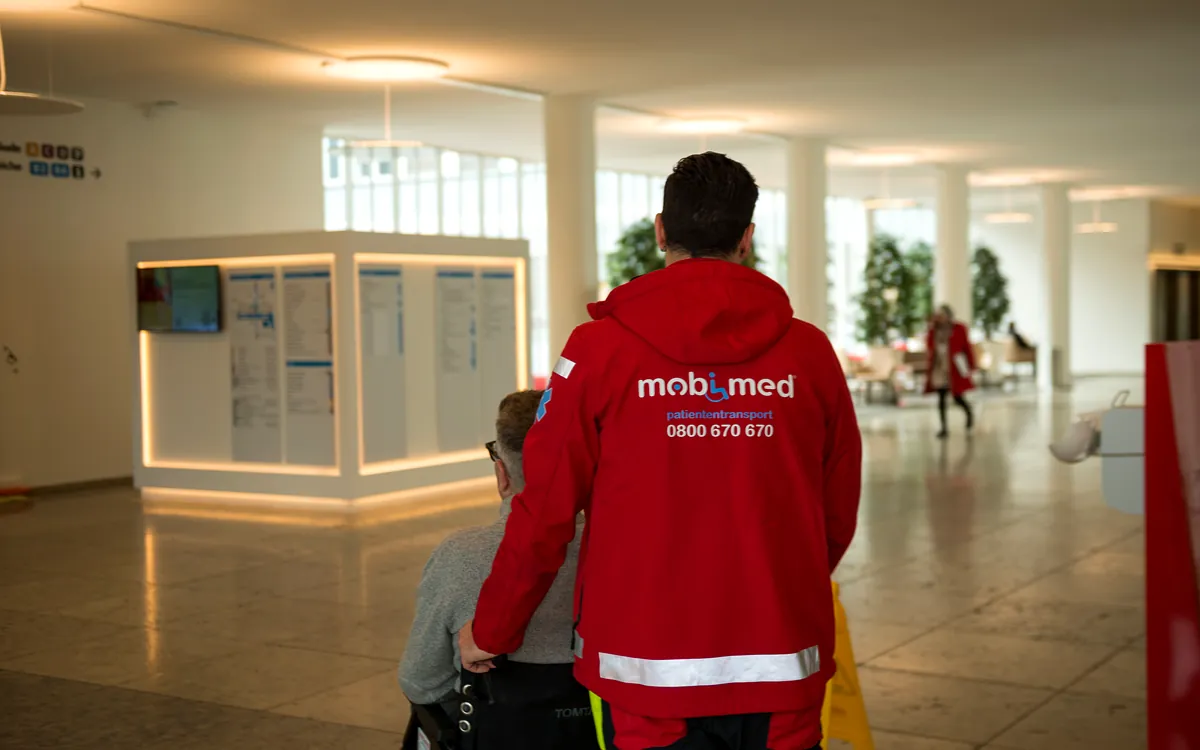 Mobimed-Mitarbeiter schiebt einen Patienten im Rollstuhl durch die Eingangshalle eines Spitals